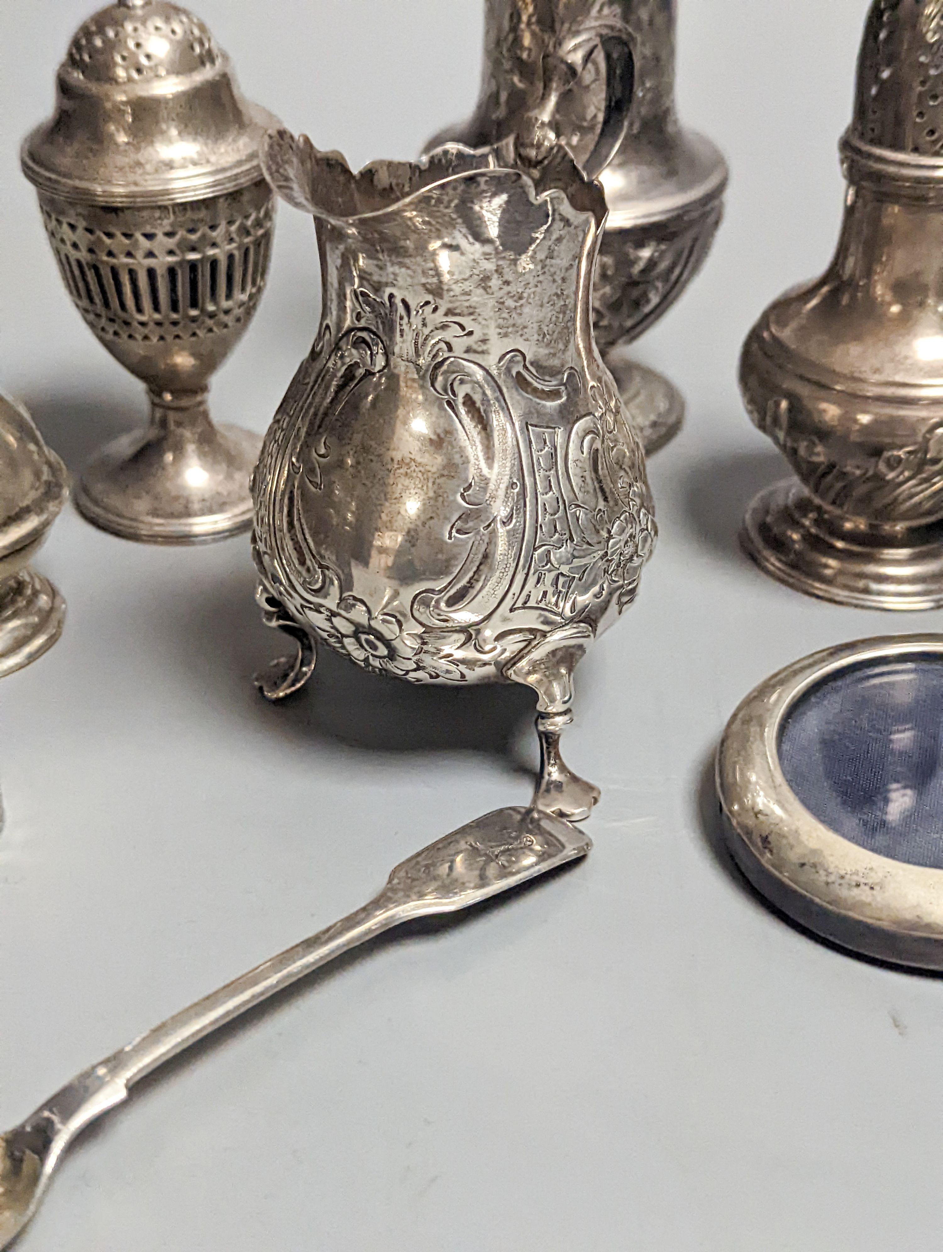 A Victorian embossed silver cream jug, London, 1883, 92mm, two 18th century silver pepperettes, one with plated cover, four later condiments (two silver), a silver spoon and small 800 standard photograph frame.